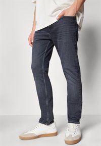 Jeans de denim azul oscuro con corte slim, que presentan una textura suave, diseño de cinco bolsillos y sutiles acentos de costura. Combinados con zapatillas blancas.
