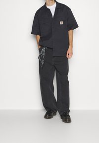Schwarzes Kurzarmhemd mit zwei Fronttaschen, kombiniert mit dunklen weiten Jeans. Ein schwarzes, gemustertes Bandana steckt in der Taschen der Jeans.