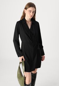 Rochie neagră, cu mâneci lungi, guler și detaliu cu fundă pe lateral. Material neted cu o siluetă clasică, asortată cu o geantă verde.