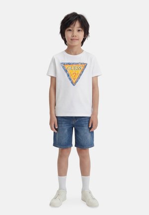 Αγόρι που φοράει λευκό κοντομάνικο μπλουζάκι με πολύχρωμο λογότυπο Guess, τζιν σορτς, λευκές κάλτσες και λευκά αθλητικά παπούτσια, στέκεται στραμμένο προς τα εμπρός.