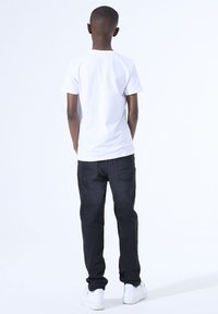 Witte t-shirt met korte mouwen, zwarte spijkerbroek en witte sneakers; gezien van achteren tegen een effen witte achtergrond.