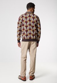 Il maglione presenta un audace motivo foglia multicolore in tonalità di bordeaux, giallo e bianco, con orlo a coste. Pantaloni beige e scarpe con motivo.