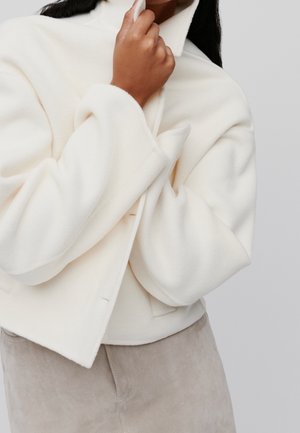 Personne portant une veste douce couleur crème avec un col montant, tenant le col près de son visage, associée à un pantalon beige clair.