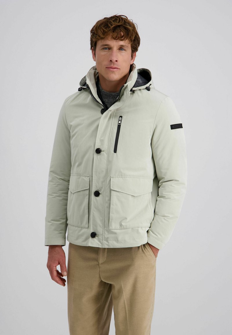 Man in een lichtgrijze hooded jacket met zwarte knopen en een ritszak op de borst, gecombineerd met beige broek, staand tegen een eenvoudige achtergrond.
