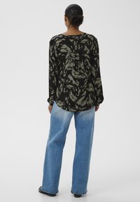 Femme aux cheveux foncés attachés en chignon, portant un chemisier à manches longues noir et gris à motifs et un jean large bleu clair, de dos.