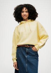 Ung kvinne med krøllete hår som har på seg en gul hettegenser og et high-waisted jeansminiskjørt med brunt belte, med den ene hånden i lomma.