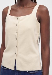Femme portant un top en maille beige sans manches avec boutons, associé à un jean bleu foncé, présenté des épaules aux hanches sur un fond uni.