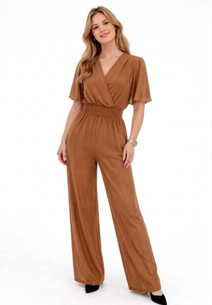 Kobieta o długich blond włosach, ubrana w brązowy kombinezon z dekoltem w kształcie litery V, szerokimi nogawkami i falującymi rękawami, stojąca na białym tle.