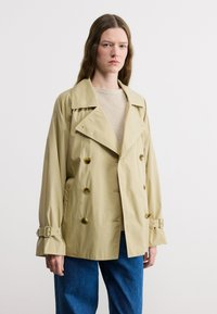 Jeune femme aux longs cheveux portant un trench-coat beige à double boutonnage et un jean bleu, sur un fond blanc uni.