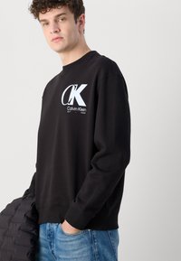 Hanorac negru din bumbac, cu un logo alb "CK" și "Calvin Klein EST 1968" pe față. Mâneci lungi, manșete și tiv cu dungi.