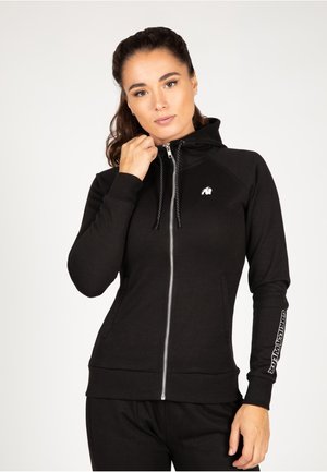 Zwarte rits hoodie met een gestructureerde afwerking, aansluitend ontwerp en koordjes. Heeft een klein logo op de borst en bedrukte tekst op de mouw.