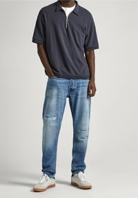 Mörkgrå pikétröja med krage och dragkedja framtill, kombinerad med ljusblå slitna jeans och vita sneakers med bruna sulor.