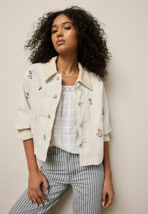 Frau mit lockigem Haar, die eine cremefarbene, mit Blumen bestickte Jacke, eine weiße Spitzenbluse und blau gestreifte Hosen trägt und vor einer schlichten Wand steht.