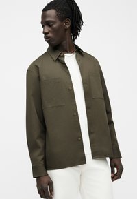 Άνδρας με σκουρόχρωμο δέρμα και dreadlocks που φορά ένα ανοιχτό ελαιχί πράσινο πουκάμισο πάνω από λευκό t-shirt και λευκό παντελόνι, κοιτάζει προς τα αριστερά του.