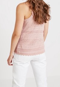 Světlý růžový háčkovaný top s volnými vzory a ozdobným zakončením ve tvaru vlnky, doplněný bílými džínami. Důraz na texturu a design.