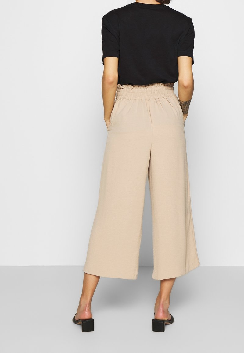Femme portant un pantalon court évasé beige avec une taille élastique et un haut noir à manches courtes, se tenant avec les mains dans les poches, vue de dos.