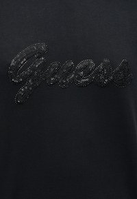 Μαύρο T-shirt με λογότυπο "Guess" διακοσμημένο με μαύρες στρασ πέτρες, δημιουργώντας μια υφή με λαμπερό αποτέλεσμα. Λείο ύφασμα με εφαρμοστή σχεδίαση.