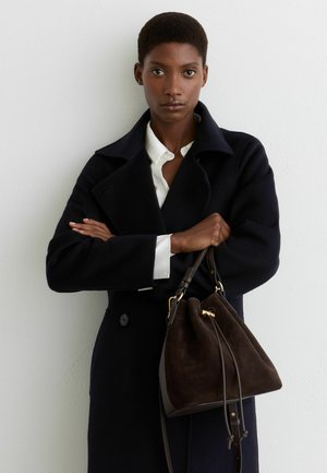 Žena s krátkymi vlasmi, ktorá má na sebe tmavý kabát cez bielu košeľu, drží tmavohnedú semišovú kabelku so zlatými kovovými doplnkami pred obyčajnou stenou.