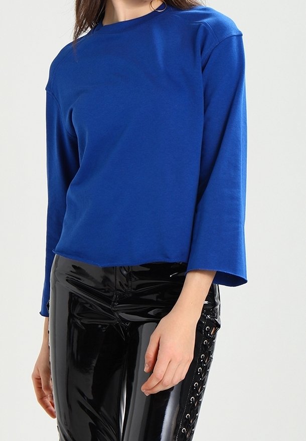 Femme portant un haut bleu vif à manches longues et un pantalon noir brillant avec des détails à lacets sur les côtés, debout devant un fond neutre.