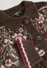 Bruine gebreide cardigan met roze en groene bloemenpatronen, zwarte knopen, geribde kraag en mouwen, gelabeld NEXT, close-up beeld.