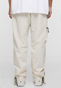 Pantaloni impermeabili color crema con vestibilità rilassata. Dettagli notevoli includono una tasca laterale con zip e chiusure regolabili alle caviglie.