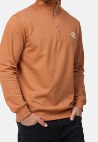 Homme portant un pull orange à manches longues avec une fermeture éclair au quart, des poignets côtelés et un petit logo blanc sur le côté gauche de la poitrine, mains dans les poches d’un pantalon foncé.