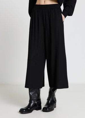 Schwarze, weit geschnittene, kurze Hose mit elastischem Bund, kombiniert mit schwarzen Lederstiefeln bis zur Mitte der Wade an einer Person mit einer Hand in der Tasche.