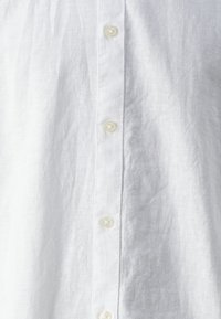 Camisa de lino blanca con botonadura frontal vertical, tela texturizada y acabado semitransparente, con botones de plástico transparente y diseño minimalista.