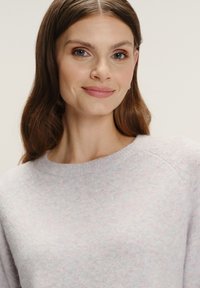 Kobieta z długimi brązowymi włosami, nosząca jasnofioletowy sweter typu crewneck, delikatnie uśmiechająca się na jednolitym jasnym tle.
