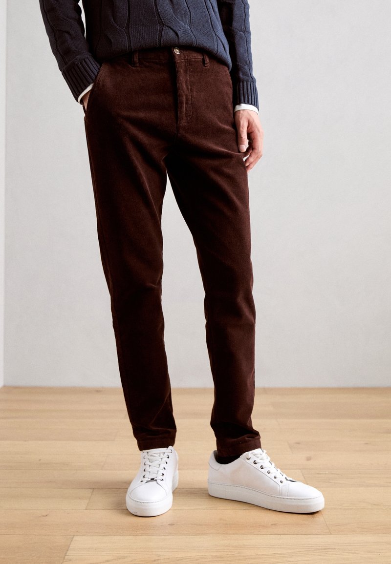 Bruna kordbyxor med en avsmalnande passform, med en mjuk yta och subtila linjer. Kombinationen med vita sneakers och en mörk tröja.