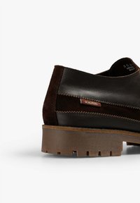 Zapato de cuero marrón con acentos de ante. Cuenta con una suela robusta, detalles cosidos y una pequeña etiqueta con el logo de "SCALPERS".