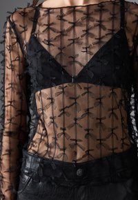 Top trasparente nero con ricami floreali, caratterizzato da scollatura quadrata e maniche lunghe sopra un bralette a triangolo nero.