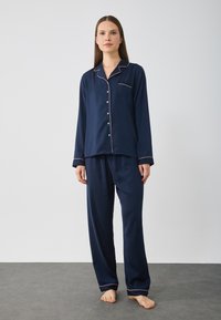 Mörkblå pyjamasset med lång ärm och byxor. Har en kontrasterande rosa kant, en framficka och knäppning med knappar. Slät tyg.