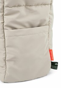 Borsa tote imbottita beige con una texture morbida, dotata di una tasca laterale e etichette che indicano materiali ecologici. Forma compatta e rettangolare.