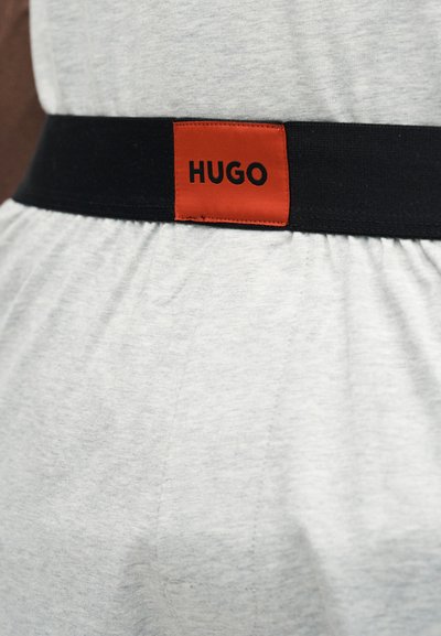 Ceinture élastique noire avec une étiquette rouge portant le mot « HUGO » en noir, portée sur un tissu gris clair.