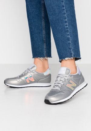 Füße mit silbernen New Balance 500 Sneakern kombiniert mit blauen Jeans mit ausgefranstem Saum auf einem einfarbigen hellen Hintergrund.