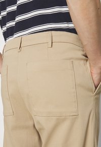 Pantalon en mélange de coton couleur sable, coupe droite, avec deux poches arrière et passants pour ceinture, associé à une chemise rayée bleu marine et blanc.