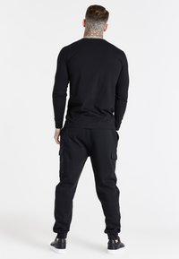 Musta pitkähihaine paita ja yhteensopivat collegehousut. Kankaan näennäinen pehmeä pinta, yksinkertainen muotoilu ja näkyvät cargo-taskut housuissa.