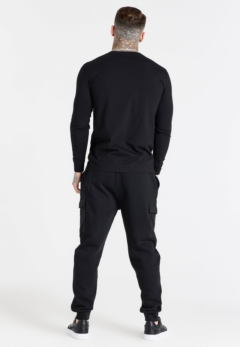 Musta pitkähihaine paita ja yhteensopivat collegehousut. Kankaan näennäinen pehmeä pinta, yksinkertainen muotoilu ja näkyvät cargo-taskut housuissa.