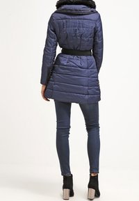 Chaqueta de plumas en color navy con cintura ajustada, cuello de piel sintética y textura acolchada. Llevada sobre jeans oscuros ajustados y botines negros de tacón.