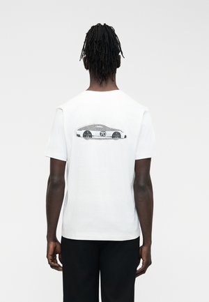 Sötét bőrű férfi dreadlock hajjal, fehér pólóban, amelyen fekete-fehér sportautó grafika és a 70-es szám látható a hátoldalon, fekete nadrágban.