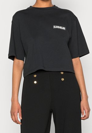 Persoon die een zwarte cropped T-shirt draagt met een klein wit logo en een zwarte hoge taille broek met vier gouden knopen aan de voorkant.