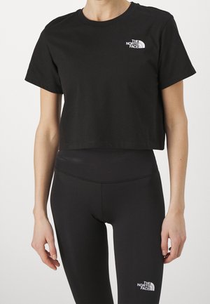 Frau trägt ein schwarzes kurzes T-Shirt und Leggings, beide mit weißem The North Face-Logo, steht vor einem einfarbigen hellen Hintergrund.