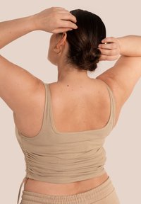 Femme aux cheveux foncés attachés en chignon, portant un débardeur côtelé beige et un pantalon assorti, ajustant ses cheveux avec les deux mains derrière la tête.