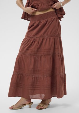 Femme portant une jupe longue marron à étages, soulevant légèrement le haut assorti, associée à des sandales beiges à talons bas et à bouts ouverts.