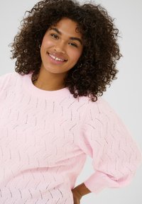 Pull en tricot rose avec une coupe ample, un motif respirant et des poignets côtelés. La texture présente un design subtil en vagues.