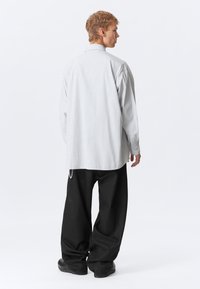Jovem em pé, de lado, vestindo calças pretas largas, uma camisa oversized a riscas brancas e sapatos pretos, em fundo branco.