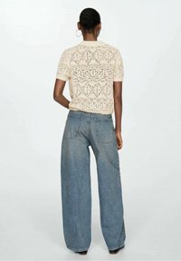 Haut beige en maille avec manches courtes et motif texturé, associé à un jean en denim bleu ample. La tenue est vue de dos.