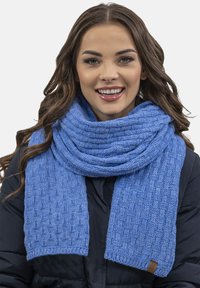 Fular bleu tricotat, cu un model texturat, țesătură groasă și o etichetă accent. Învelit în jurul gâtului, o parte a unei jachete puffer este vizibilă.