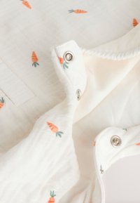 Vêtement blanc doux pour bébé avec de petites imprimés carottes orange et des boutons-pression métalliques fixant les couches de tissu.
